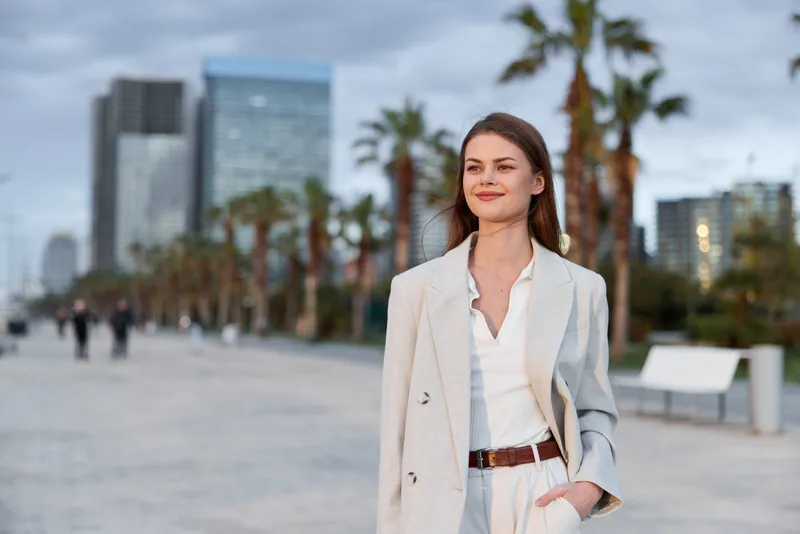 Confident woman in beige coat walking outdoors in urban business district with palm trees and skyscrapers in background