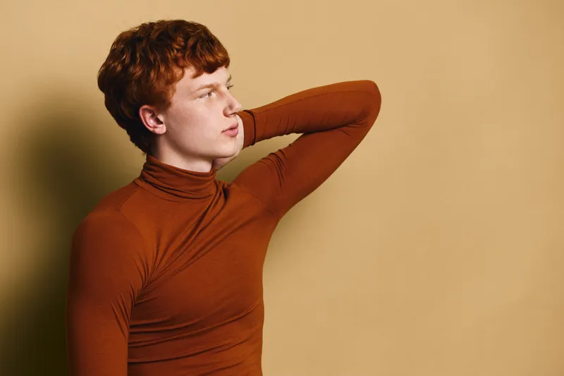 Redhead man in brown turtleneck poses with arm behind head against beige wall, looking thoughtful and relaxed in casual studio portrait with soft lighting and neutral colors.
