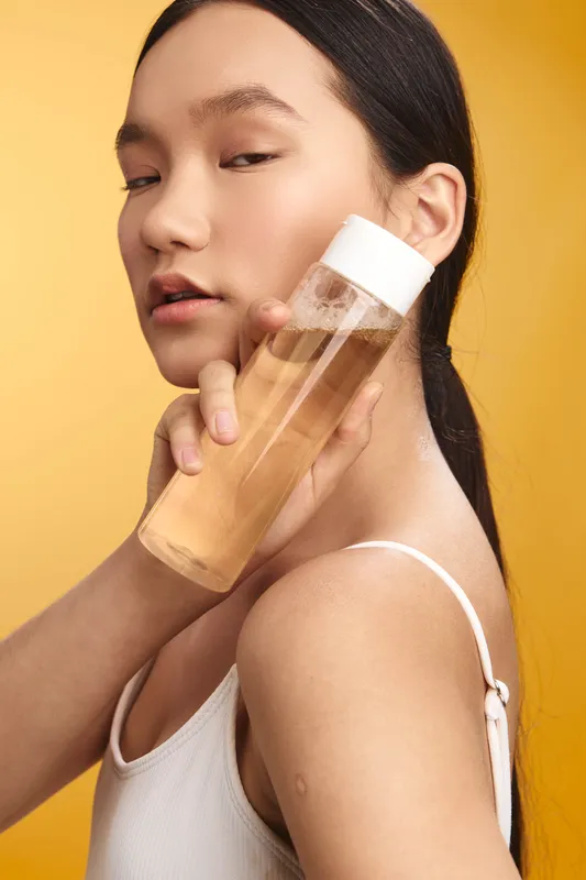 asian woman holds a clear skincare bottle against a soft studio backdrop, a calm portrait highlighting a simple beauty routine and clean, minimalist aesthetic.