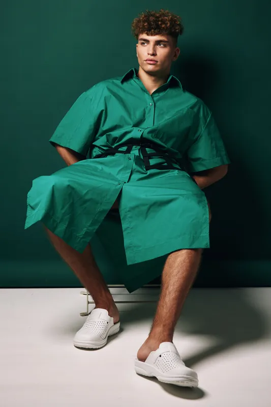 Young man sitting with hands behind back against dark green background wearing a green short sleeve buttoned dress and white slip-on shoes at studio indoor.