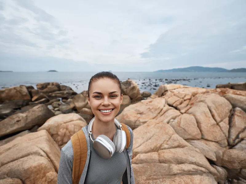 Happy young woman with headphones on rocky beach, enjoying a tranquil summer day by the sea, showcasing a relaxed and cheerful outdoor lifestyle