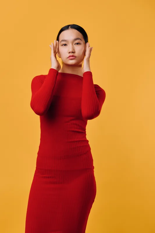 Woman in red dress standing and touching face with hands posing on orange background. Fashion model with long black hair and confident expression in studio lighting.