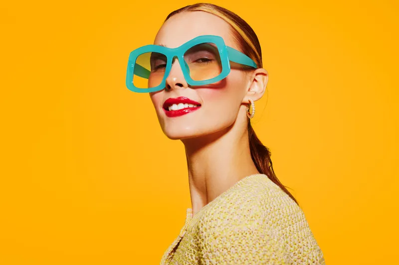 Stylish woman with bright blue oversized glasses and red lipstick poses confidently against vibrant yellow background. Concept of fashion, beauty, and modern eyewear.