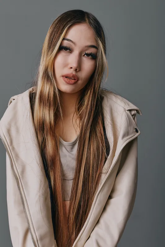 Young woman with long straight hair wearing light jacket and apron posing in studio. Fashion model with natural makeup in casual outfit on neutral background with serious expression.