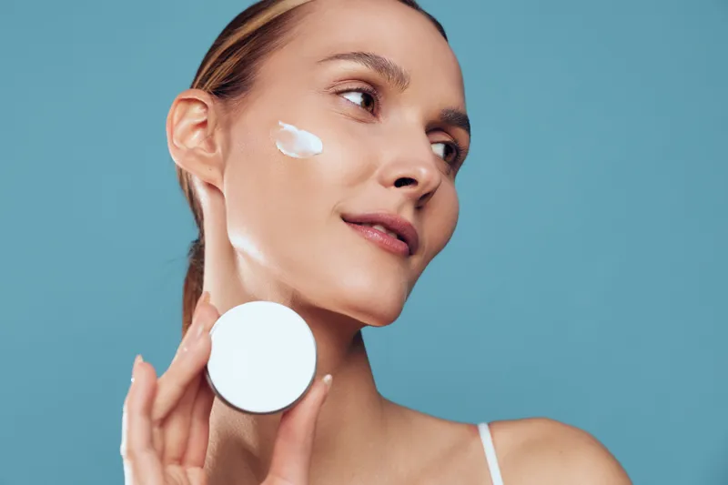 Closeup of a woman applying white cream to her cheek, holding a round container against a blue background. Focus on skincare, wellness, and self care with soft studio lighting.
