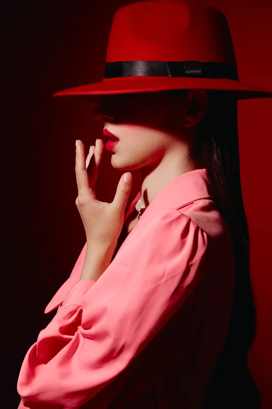 Stylish young woman wears a wide red hat and bright pink blouse posing in dramatic shadow. The vibrant colors create a striking beauty and fashion concept.