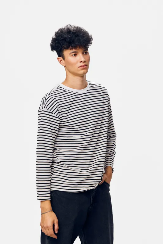 Young man wearing a striped long sleeve shirt and black pants standing against a white background, looking thoughtful with casual style and curly hair.