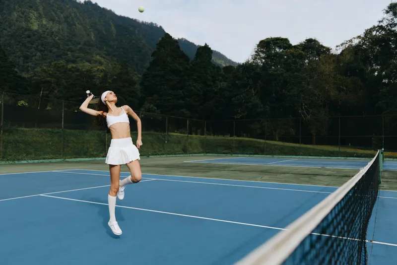 Female tennis player in white athletic wear serving a tennis ball on a blue court surrounded by green trees, showcasing action and determination for sports enthusiasts