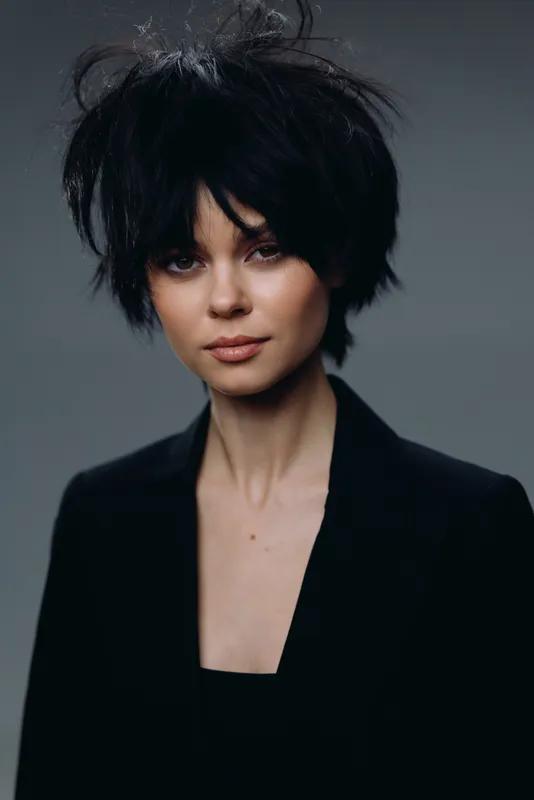 Portrait of a young woman with dark messy hair wearing a black blazer and neutral expression against a gray background.