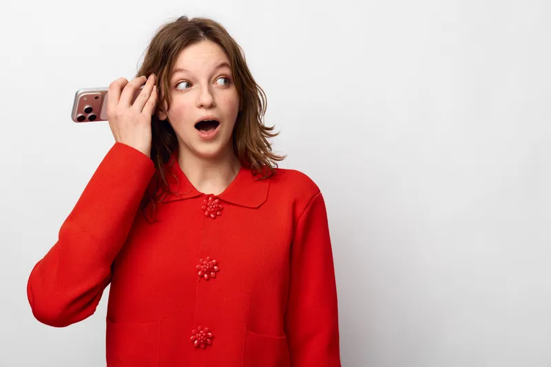 Surprised young woman in red sweater holds smartphone to ear, looking away with open mouth. Concept of unexpected news, communication, and emotional reaction in casual indoor setting.