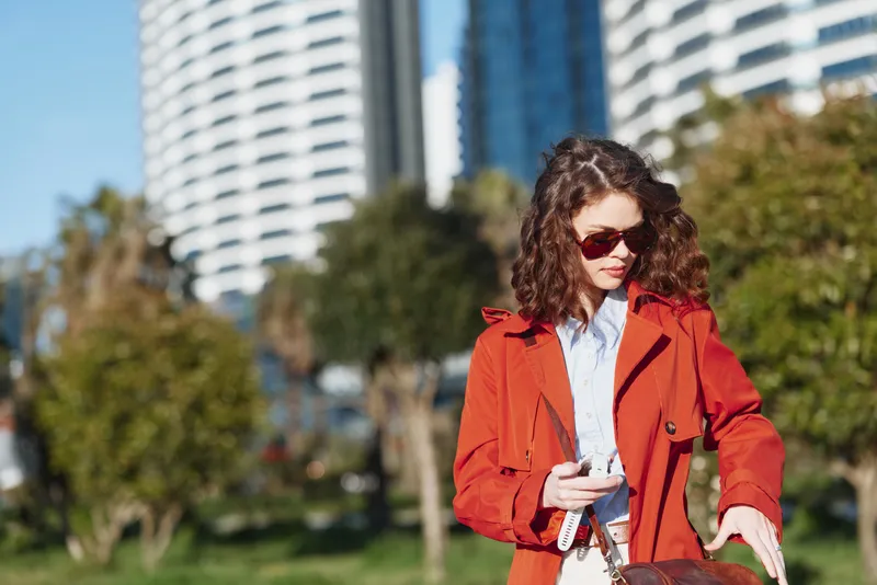 Young woman with curly hair wearing a red coat and sunglasses checks her phone while standing outdoors in an urban park on a sunny day.