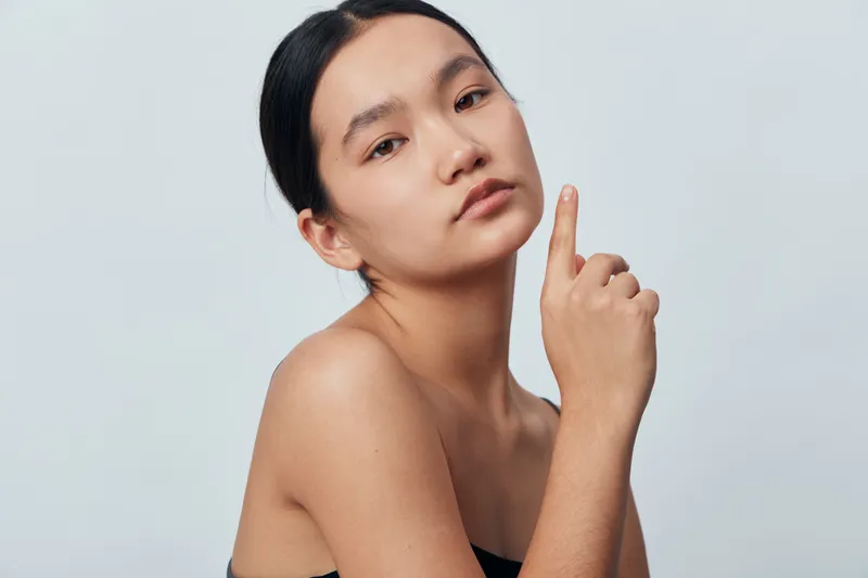 Woman with natural makeup and smooth skin posing with hand near face on light background. Beauty, skincare and portrait of female with dark hair and serious expression in studio.