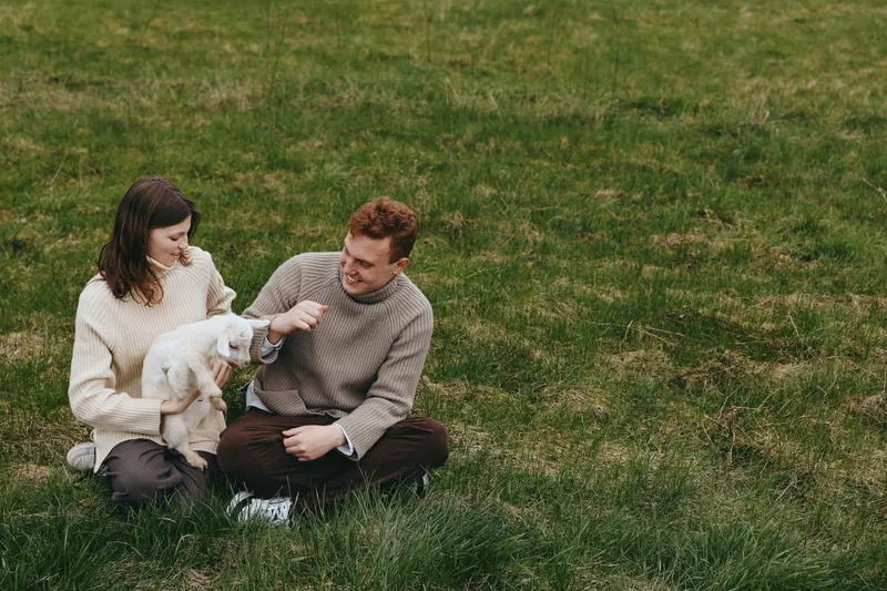 Lifestyle couple with a goat kid on a green lawn, sharing a gentle moment and laughter. Natural, candid composition emphasizes affection, outdoors living, and simple countryside joy.