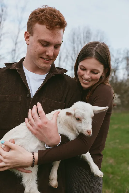Loving couple outdoors cradle a goat kid, smiling as they hold the young animal in a grassy field, warm natural light, rustic scenery, and a gentle countryside lifestyle.