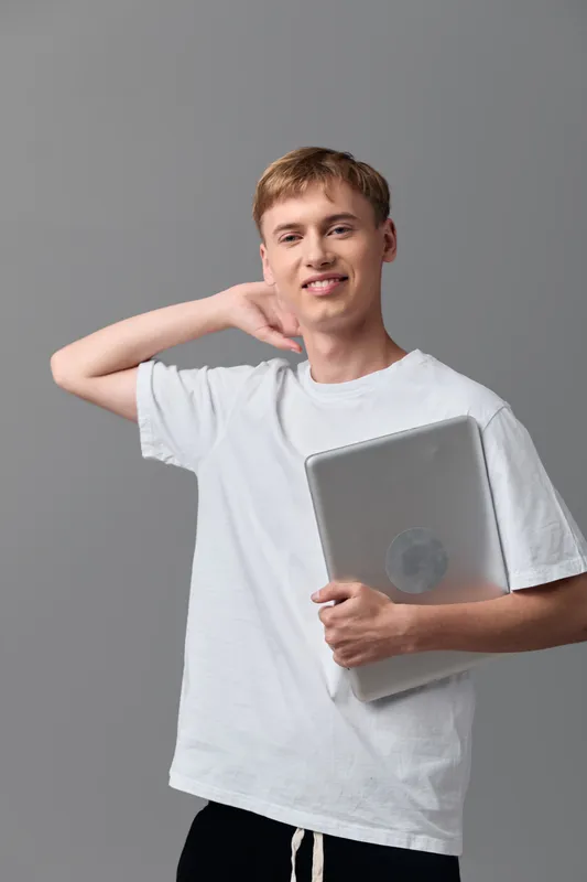 Smiling young man in a white tee holding a laptop, posing casually in a studio for education or technology themed content, showcasing relaxed learning and modern device usage