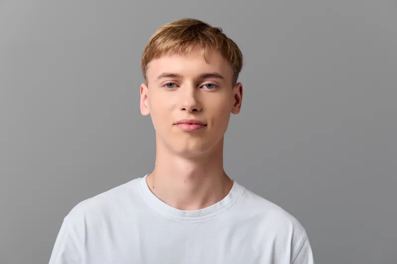 Face of a young man in a plain white tee against a neutral gray studio backdrop, calm expression, clean lighting and minimal style suitable for lifestyle, fashion and ecommerce mockups