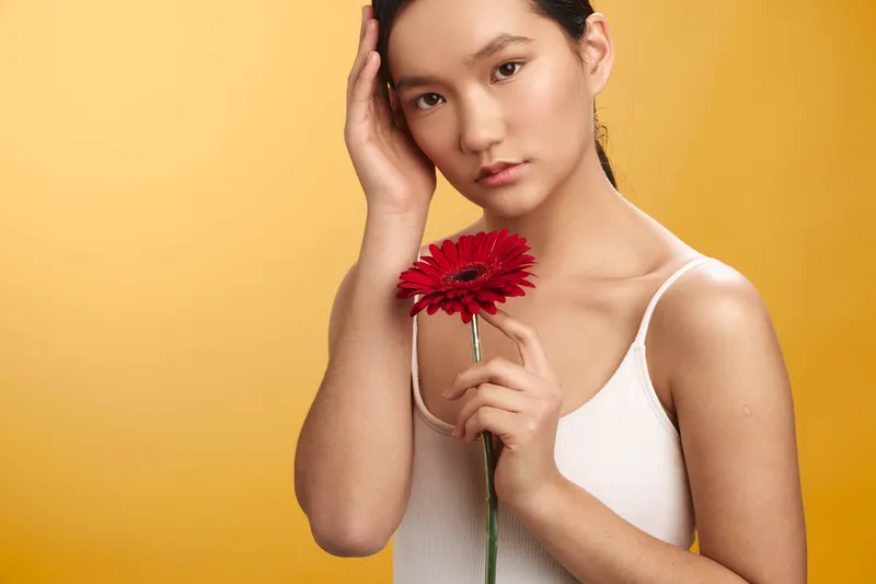 asian woman holds a red flower against a warm yellow background, presenting a calm thoughtful portrait with subtle emotion, graceful posture, and a soft harmonious composition.