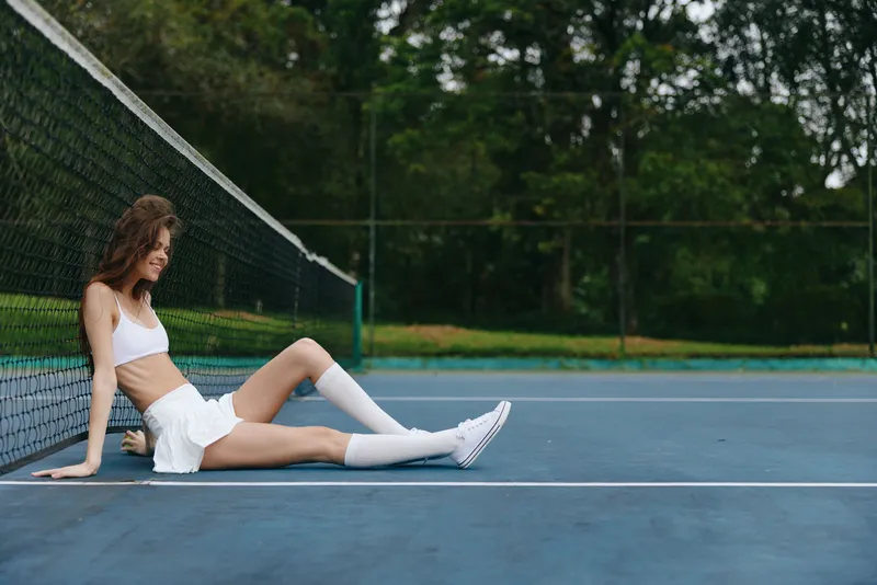 A young female tennis player in a white outfit sits on the court, displaying a relaxed expression amidst lush green surroundings, capturing a serene athletic vibe