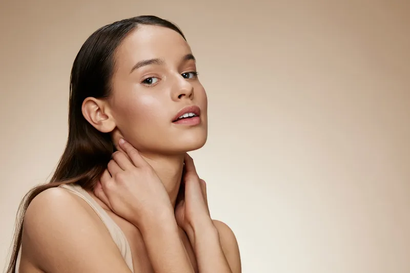 Young woman with long brown hair, natural makeup, soft expression on a beige background, showcasing beauty and skincare concepts for health and wellness