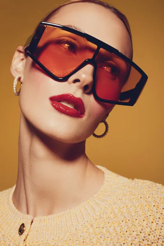 Close-up portrait of a stylish woman wearing oversized orange sunglasses and gold hoop earrings. She has red lipstick and a beige knitted sweater on a warm yellow background.