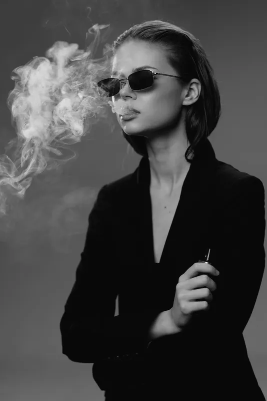 Black and white portrait of a stylish woman smoking with sunglasses in a dark setting, showcasing attitude and modern fashion.