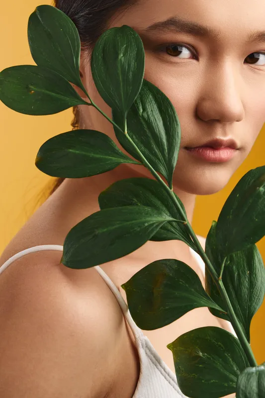 asian woman appears serene as foliage partially veils her face in a clean studio portrait, soft lighting highlights delicate skin texture and a natural botanical mood.