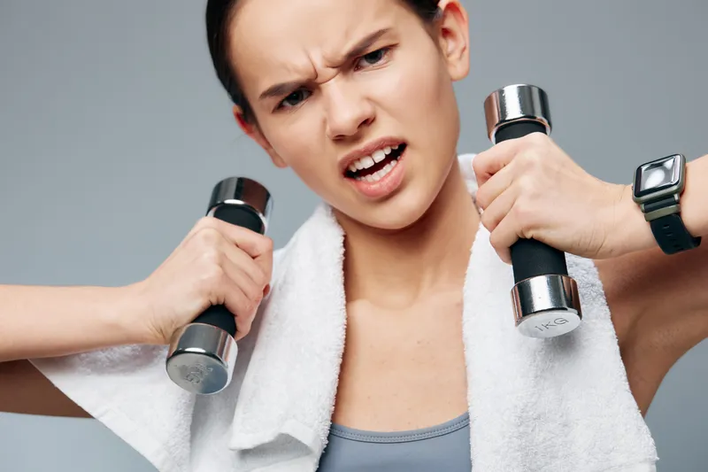 Female fitness athlete lifts dumbbells with a towel draped over her shoulders, showing intense expression during a demanding workout in a bright gym, focus on strength and form.