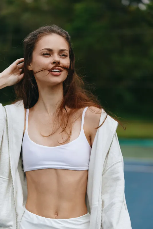 Young athletic woman smiling confidently while enjoying an outdoor workout in a white sports bra and sweatshirt, surrounded by greenery