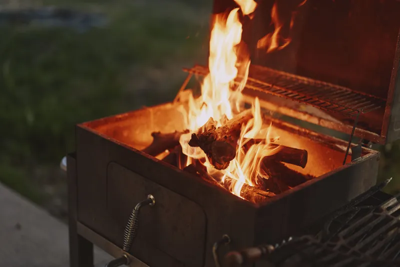 A cozy outdoor barbecue scene with flames dancing above burning wood in a grill, perfect for summer gatherings and outdoor cooking experiences
