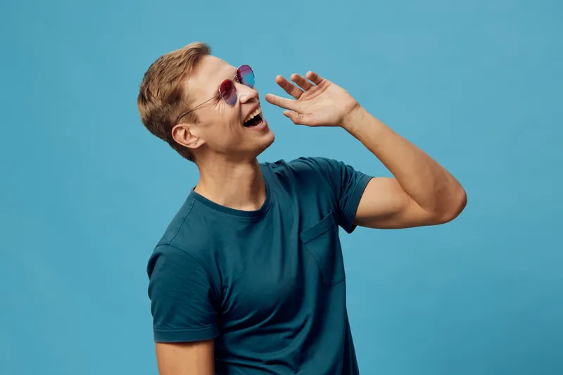 Smiling woman with short hair wearing sunglasses and blue t-shirt, expressing joy and laughter, posing against a plain blue background, studio portrait, lifestyle concept.