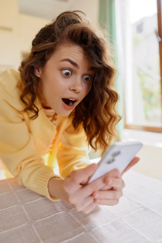 Joyful woman indoors, surprised by her phone screen, cozy home interior, soft daylight, casual outfit, candid lifestyle moment, vibrant emotions