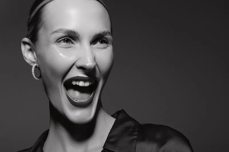 Caucasian woman with light skin laughing joyfully in black shirt and hoop earrings. Black and white portrait with dramatic lighting and expressive emotional mood.