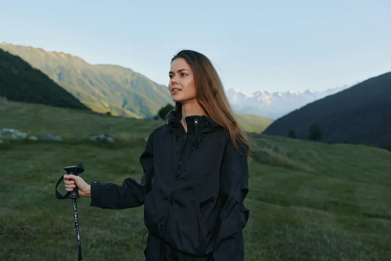 A confident hiker stands in a green valley with distant mountains, wearing a dark rain jacket and holding a trekking pole, enjoying a serene outdoor moment.