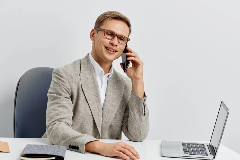 Confident smiling man in business attire talking on mobile phone while sitting at desk with laptop isolated on plain white background, professional office environment, corporate concept, success.