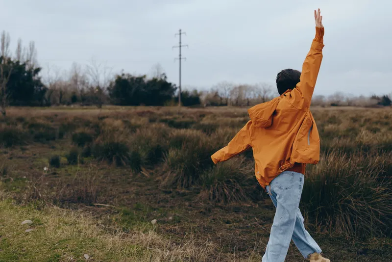 Woman lifestyle outdoors walking countryside field, orange jacket and jeans, one arm raised in cloudy air, grassy meadow background, spring calm, relaxed posture, natural colors
