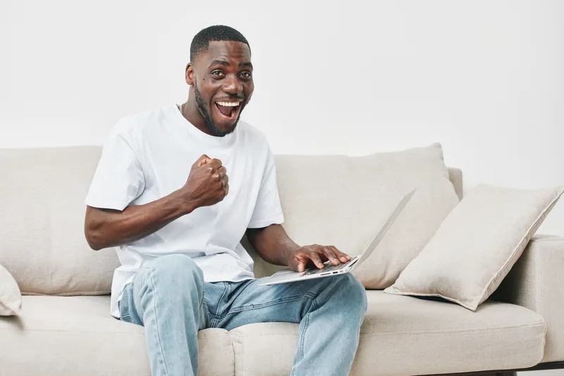 Excited young African man celebrating success while working on a laptop at home, wearing casual white t shirt and jeans in a bright living room