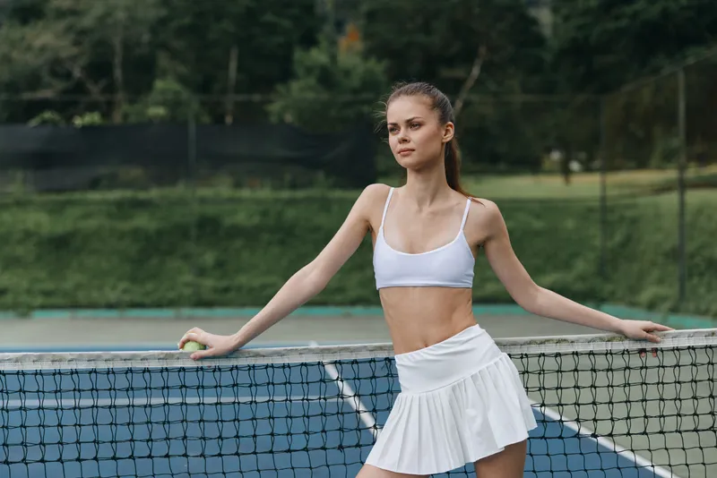 Young female tennis player in activewear poses confidently by the tennis net, showcasing her athleticism and grace, surrounded by a lush green background
