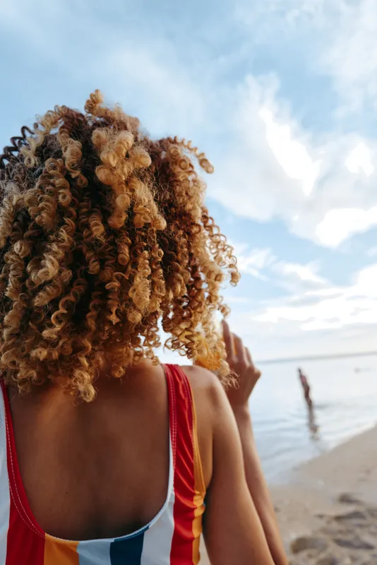 Young woman with curly blonde hair wearing a colorful swimsuit stands on the beach looking at the ocean under a cloudy sky during daytime.