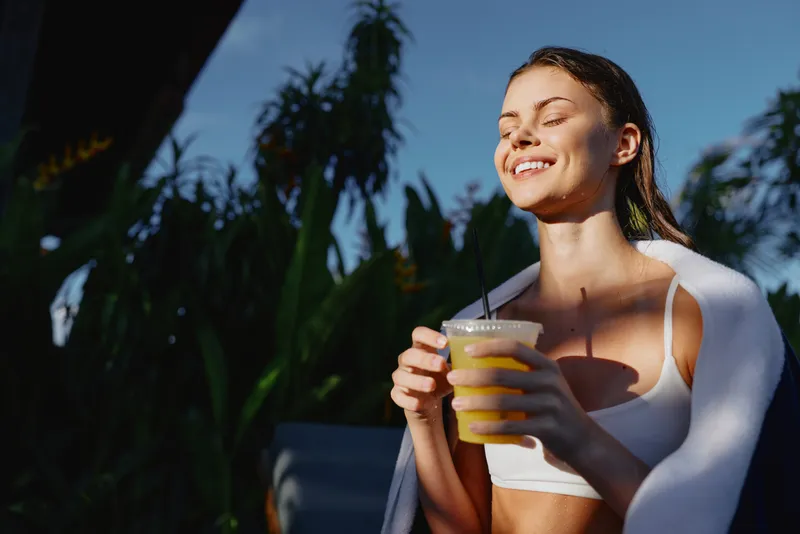 Young woman with long brunette hair enjoys a fresh juice outdoors in natural sunlight, smiling with closed eyes and wearing a white sports bra during a relaxed lifestyle moment.