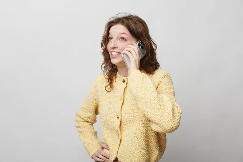 Young woman in yellow sweater talks on smartphone with smile, looking away. Casual lifestyle, communication, technology, and positive mood concept on light background.
