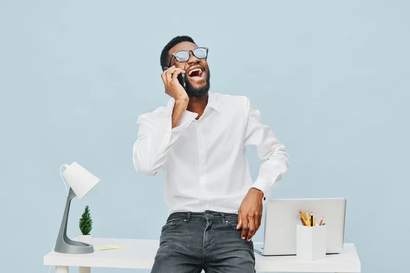 Smiling young man with beard, glasses, and white shirt talking on mobile phone at modern desk with laptop, lamp, and stationery isolated on pastel blue background, casual office lifestyle concept.