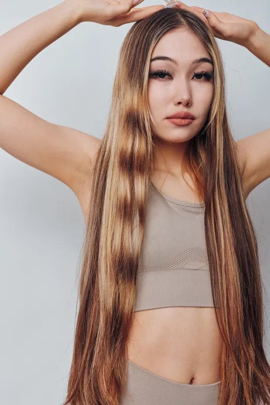 Young woman with long straight hair posing with hands above head in neutral tank top against white background for beauty and fashion concept.