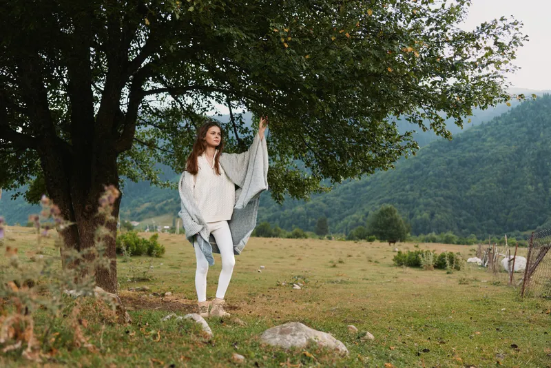 Young woman with long hair stands under a large tree in a mountainous meadow wearing a light poncho and white pants, enjoying a calm and natural outdoor moment.
