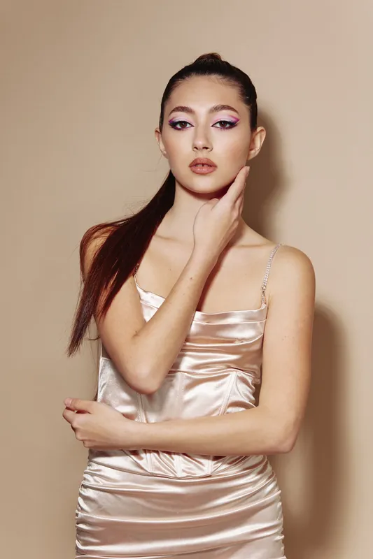 Young woman in satin dress with glossy skin and dramatic pink winged eye makeup poses against beige background. Beauty, fashion concept with creative style and elegant silhouette.