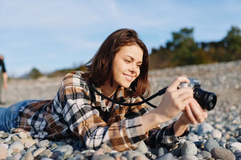 Woman photographer lying on a rocky beach, aiming a camera with a plaid shirt, casual outdoor moment, focusing on composition and natural light for creative storytelling.