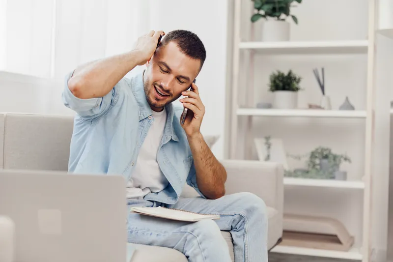 Happy man on phone, wearing casual clothing, smiling while sitting on a couch, in a modern living room filled with plants, conveying a positive work-from-home vibe.