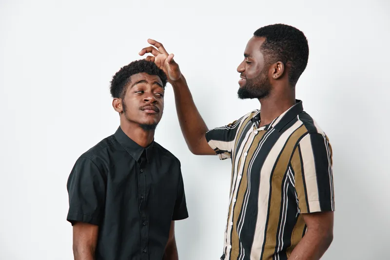 Two African American men with dark skin interacting in studio, one playfully touching the other's hair, casual clothing and friendly expression on white background.