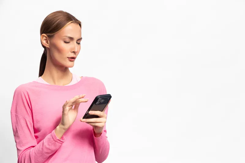 Woman in pink sweater uses smartphone with focused expression isolated on white background. Concept for mobile technology, communication, and digital interaction.