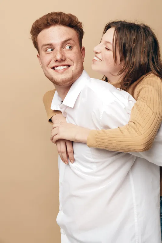 Happy young couple in casual clothes share joyful moment, woman hugging man from behind. Light beige background, smiling faces, concept of love, friendship, and togetherness.