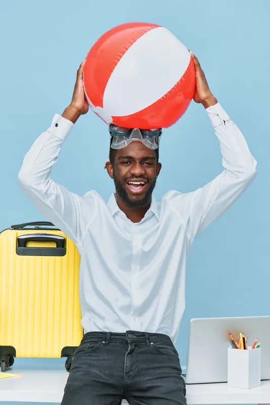 Smiling man holding large colorful inflatable beach ball above head in bright modern office setting. Casual summer vacation theme, joyful expression, casual white shirt, summer accessories, playful mo